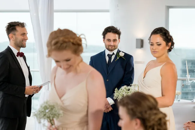 Wedding ceremony with smiling man in a blue suit and women in light dresses holding flowers.