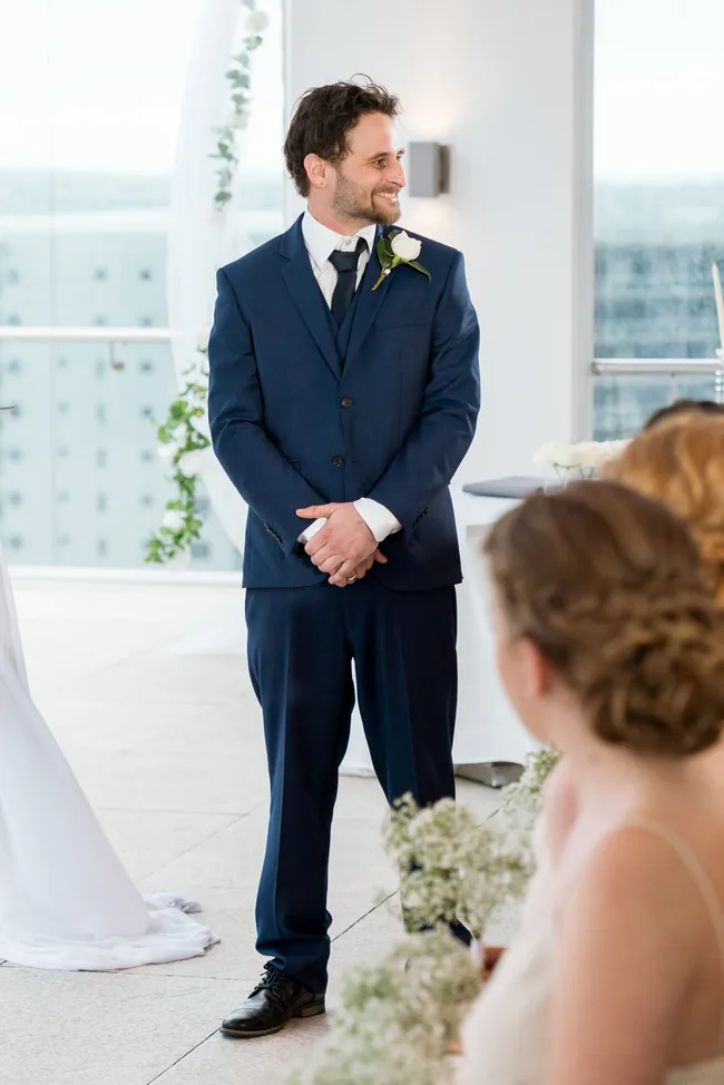 Man in blue suit with white rose boutonniere smiling at woman in foreground during wedding ceremony indoors.