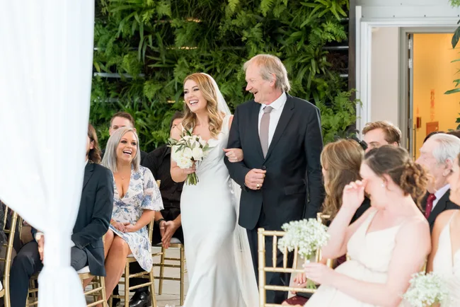 Bride in white dress walks down aisle with older man, smiling, in front of guests.