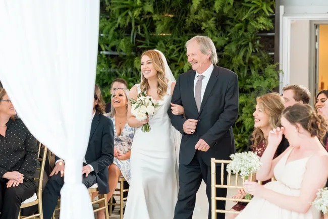 A bride in a white dress smiles while walking down the aisle with an older man at a wedding ceremony.