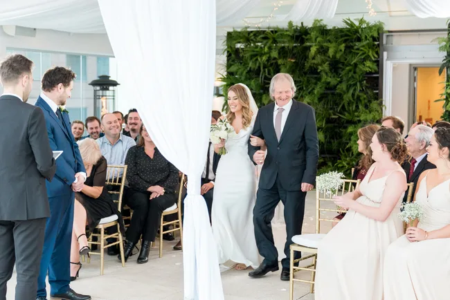 A bride walks down the aisle with an older man at a wedding, smiling guests and bridesmaids in the background.
