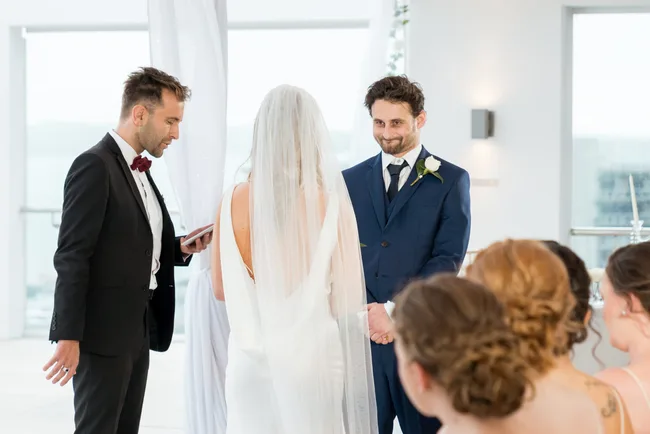 Wedding ceremony with a bride in a veil, groom in blue suit, and officiant in black suit with bow tie.