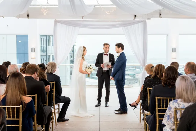 A couple stands facing each other at an indoor wedding ceremony with guests seated on both sides.