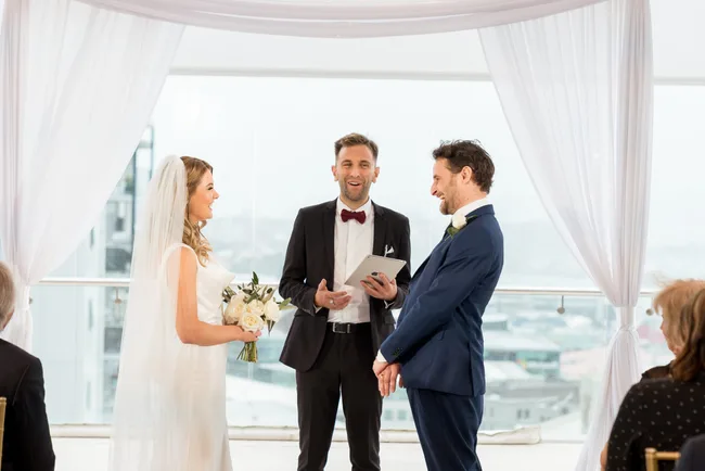 Wedding ceremony with a bride, groom, and officiant under a white draped arch, smiling during vows.