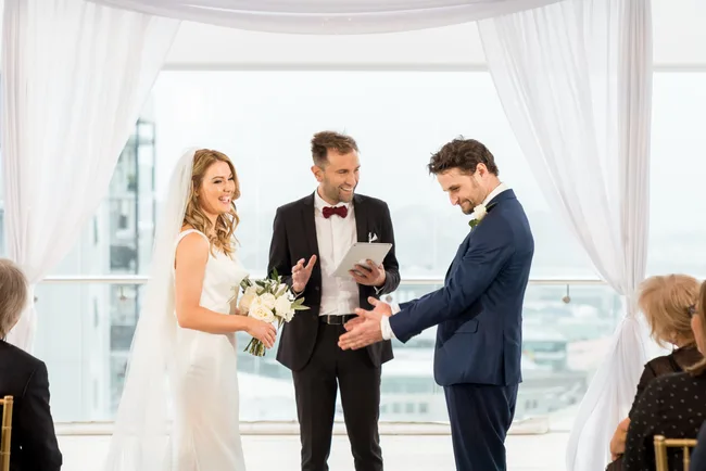 A bride and groom smile at each other during a wedding ceremony, with a joyful officiant in the background.