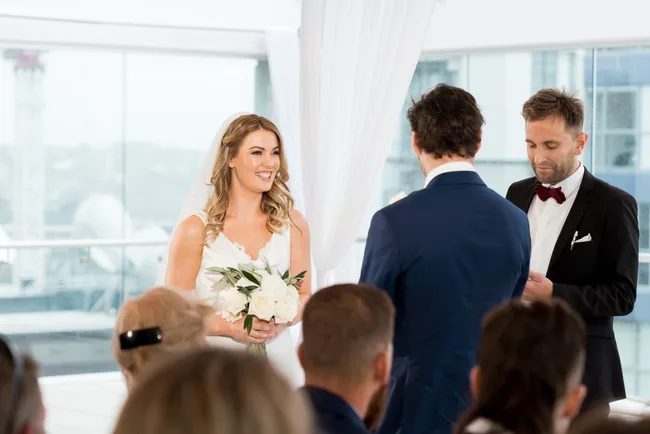 Bride smiling at groom during wedding ceremony, holding white flowers. Guests watch as officiant reads vows indoors.