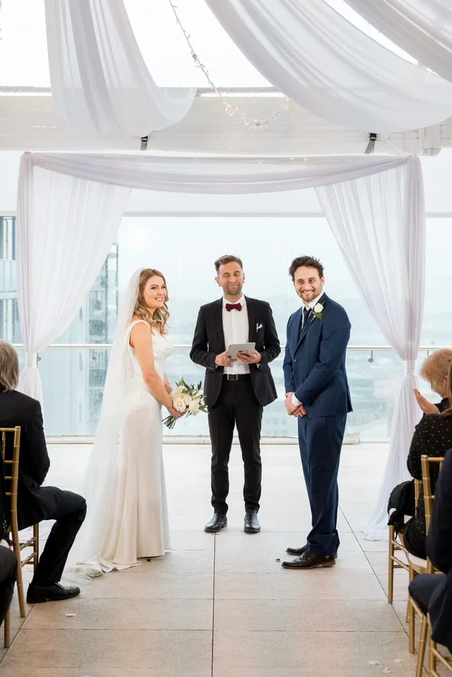 A bride and groom stand at the altar during an indoor wedding ceremony with a smiling officiant.