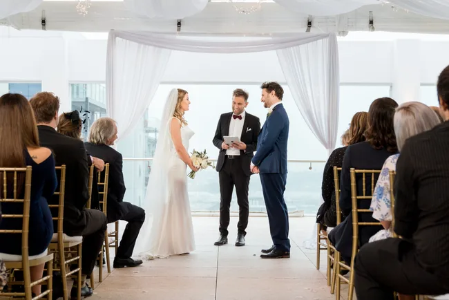 Wedding ceremony with a bride in a white dress and groom in a blue suit, officiant in the center, and guests seated.