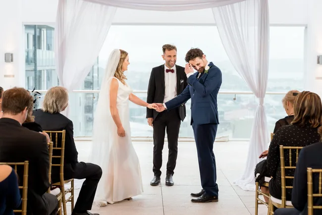 Bride and groom hold hands at indoor wedding ceremony with guests and officiant.