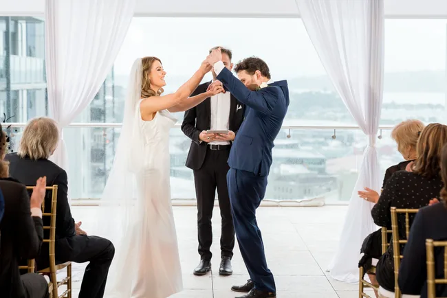A bride and groom smile and celebrate during a wedding ceremony, surrounded by seated guests.