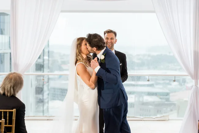 Couple kissing at wedding ceremony with officiant smiling in the background.