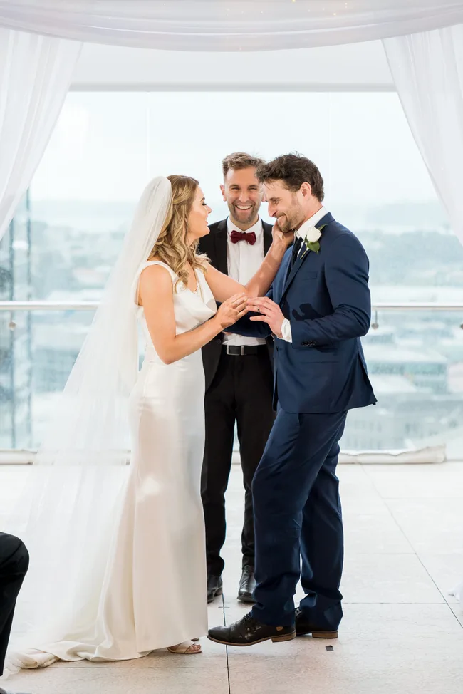 Bride and groom smiling at each other during wedding ceremony with officiant in the background.
