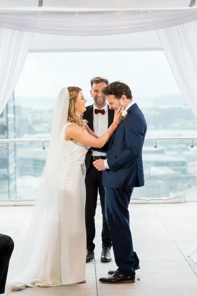 A bride and groom smiling at each other during their wedding ceremony, with the officiant in the background.