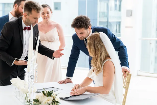 Bride signing a document at a wedding ceremony with groom and witnesses nearby in a bright room.