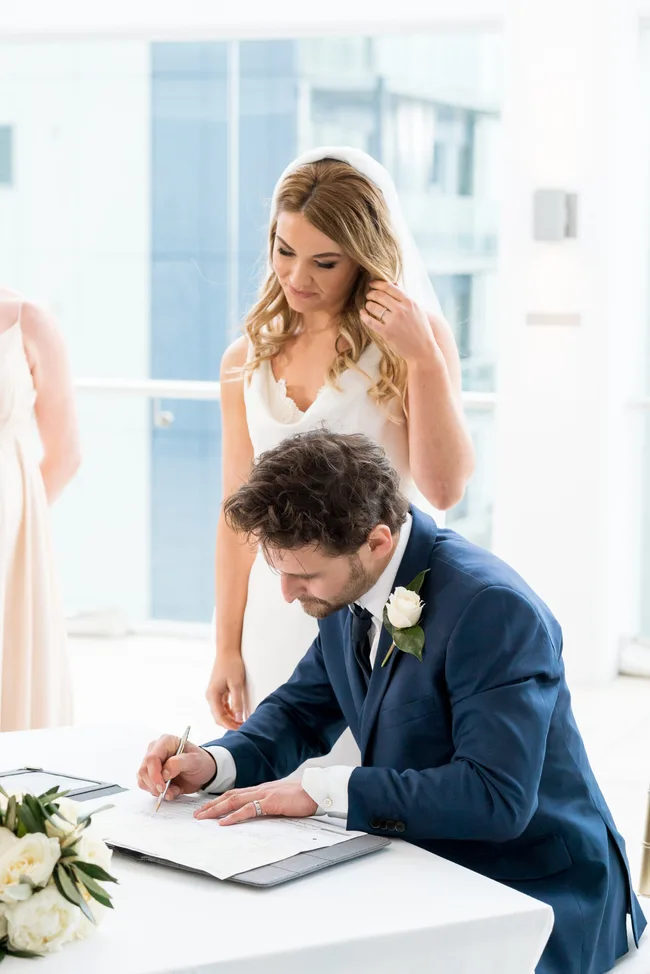 Bride stands beside groom signing a marriage certificate at a table with a bouquet, in a well-lit room.