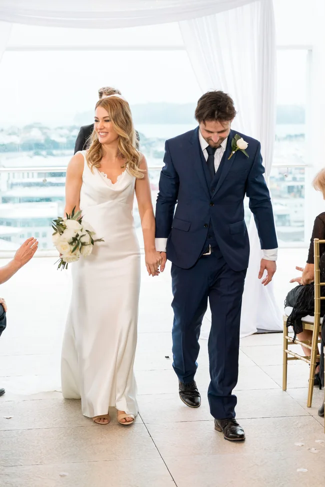 Bride in white dress and groom in blue suit walking hand in hand down the aisle at a wedding ceremony.