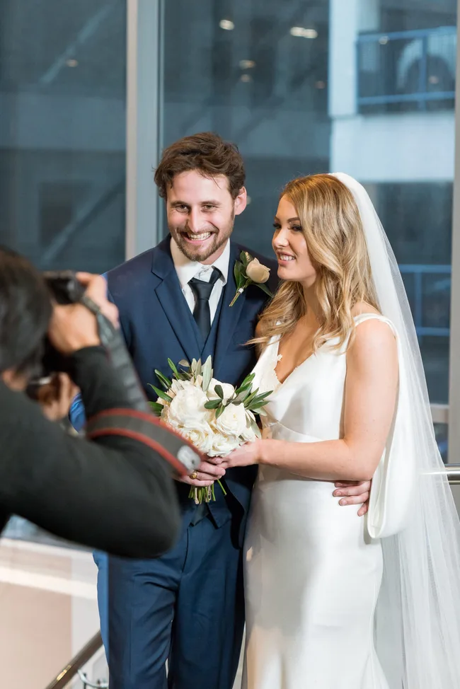 Wedding couple posing for a photo; bride in white dress with bouquet and groom in blue suit.