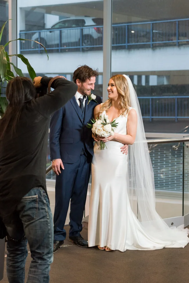 Photographer captures a smiling bride and groom. Bride in a white dress holding flowers; groom in a navy suit.