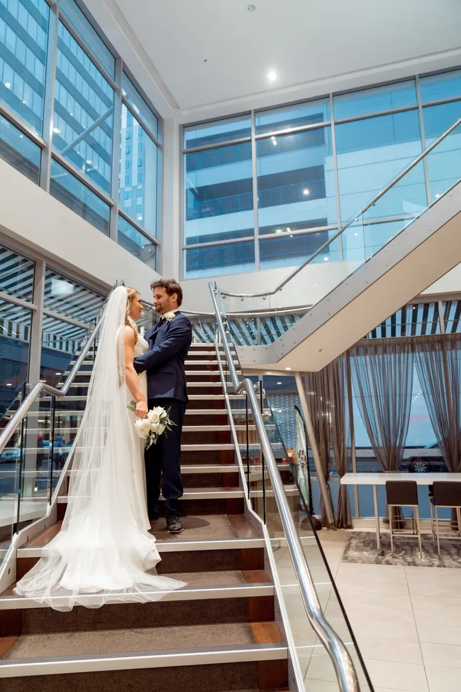 A bride and groom standing on a staircase in a modern building with large windows. Bride holds a bouquet.