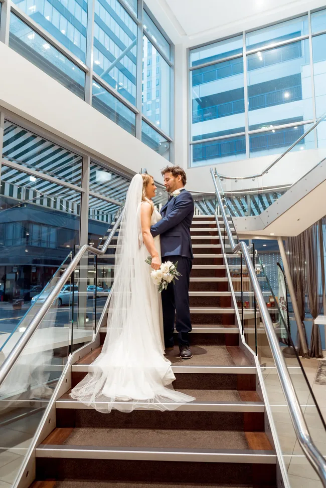 A bride and groom stand on a staircase inside a modern building, embracing, with a cityscape visible through large windows.