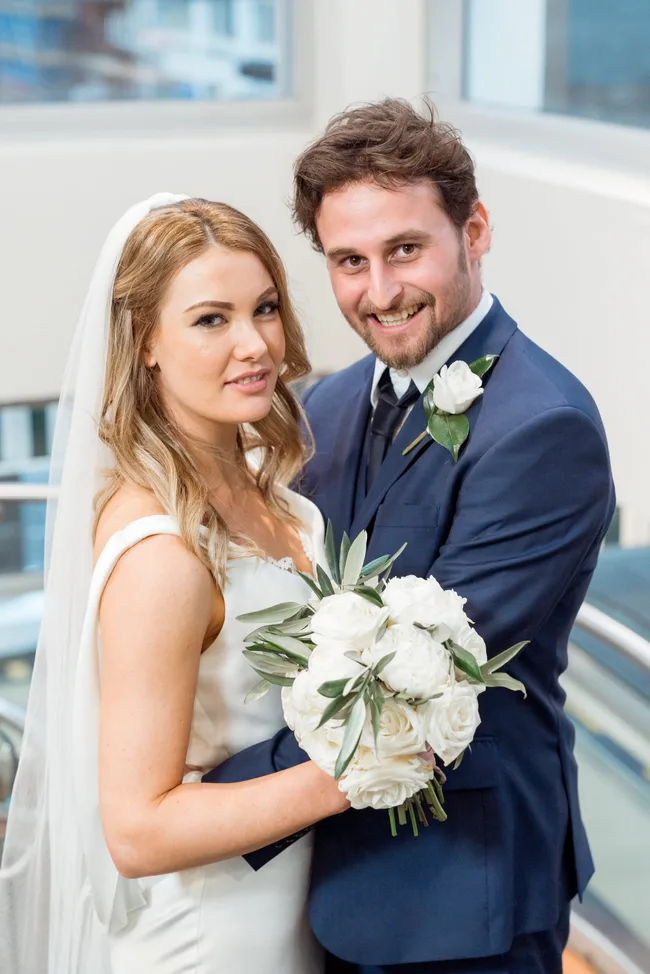 A bride and groom smiling, with the bride holding a bouquet of white roses, in a bright indoor setting.