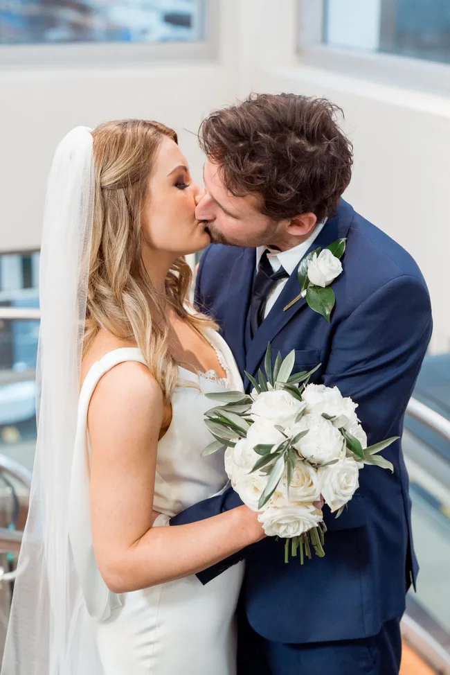Bride and groom kissing, bride holds bouquet of white roses, indoors.