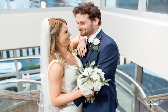 Bride and groom share a joyful moment on a staircase, both smiling and holding a bouquet of white roses.