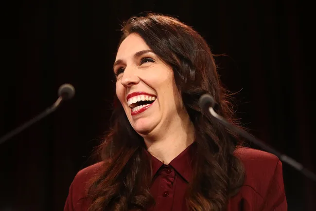 A woman with long dark hair is smiling broadly while standing at a microphone.