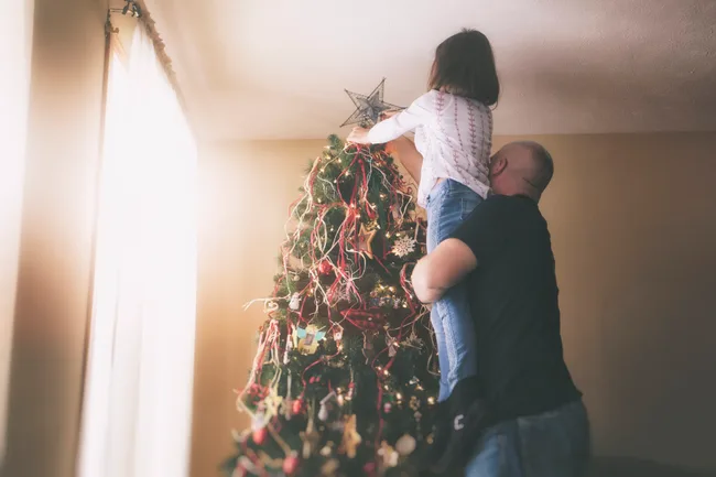 Father lifts child to place star on Christmas tree top by window light.