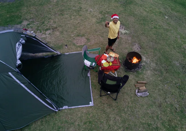 Man in Santa hat near a campfire, smiling, with tent and chairs on grass.