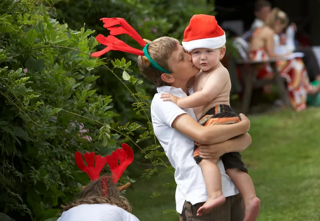Boy wearing antlers kisses a baby in a Santa hat outdoors during a festive gathering.