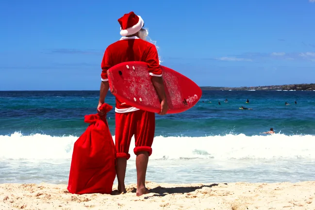 Santa in a red suit holding a surfboard and sack, standing on a sunny beach, facing the ocean waves.