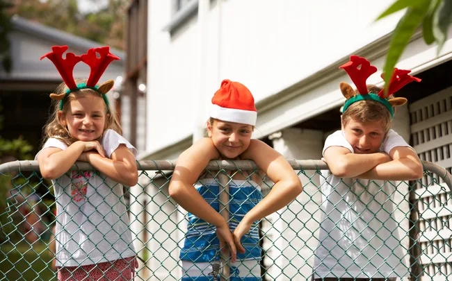 Three children leaning on a fence, wearing festive reindeer antlers and a Santa hat, smiling outdoors.