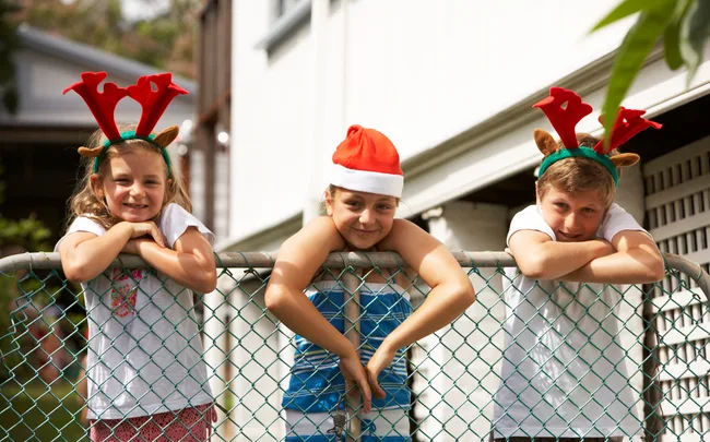 Three children leaning on a fence, wearing festive reindeer antlers and a Santa hat, smiling outdoors.