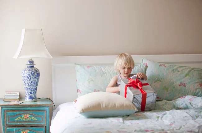 Child on bed unwrapping a gift with red ribbon, next to a blue floral lamp and teal nightstand.