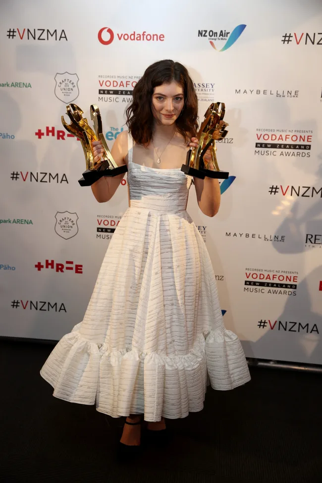 Young woman in a white dress holding two gold awards at Vodafone New Zealand Music Awards with branded background.