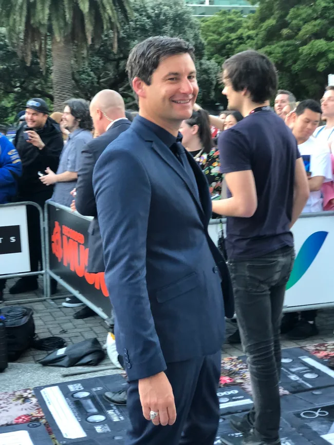 Man in a blue suit smiles while standing outdoors at an event; people and greenery in the background.