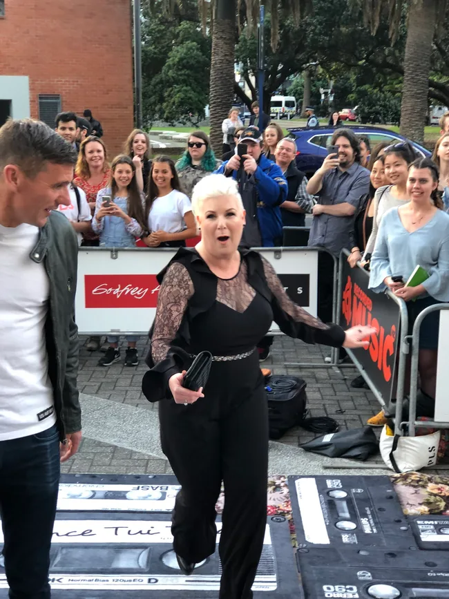 A woman in a black outfit walks down a career-themed path, smiling with people and cameras around her.