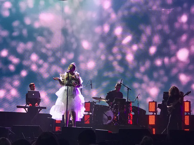 Singer performing on stage with a band, under ethereal lighting, featuring a keyboardist, drummer, and guitarist.