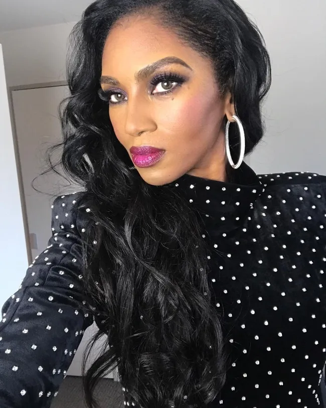 Woman with long black hair and hoop earrings, wearing a black polka-dotted top, looking towards camera.