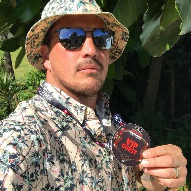 Man in floral shirt and hat holds a VIP guest badge outdoors, wearing sunglasses.