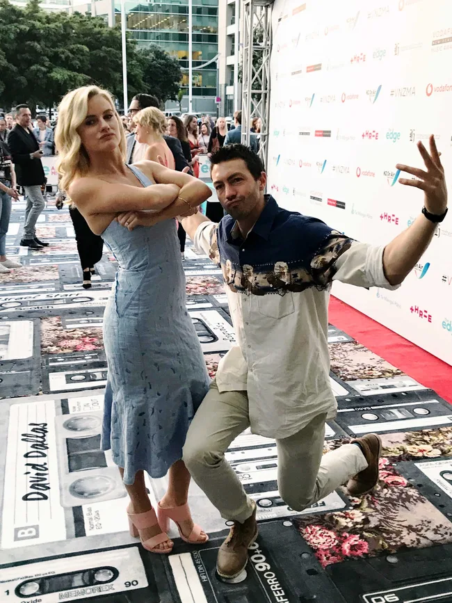 Two people pose playfully on a movie-themed carpet at an outdoor event, with a crowd and sponsor backdrop behind.