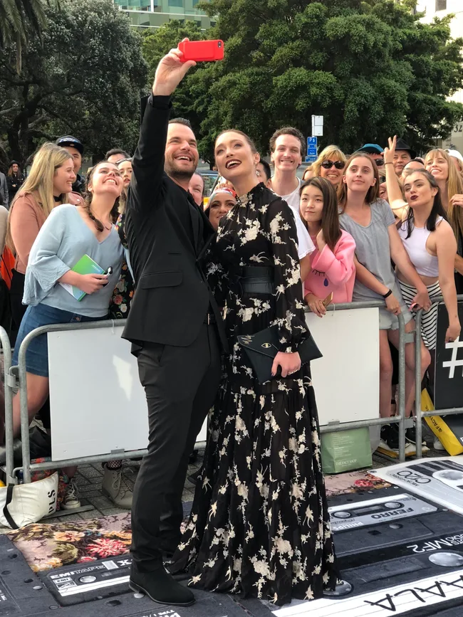 A man and woman in formal attire take a selfie with a group of smiling fans behind a barricade outdoors.