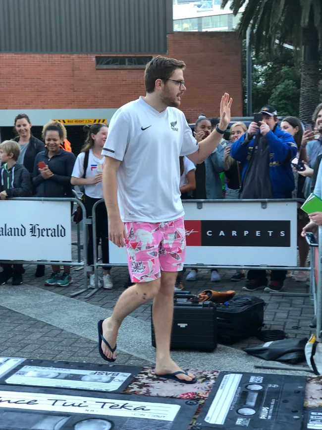 Person in a white shirt and pink shorts waves to a crowd, walking on a path with New Zealand Herald signs in the background.