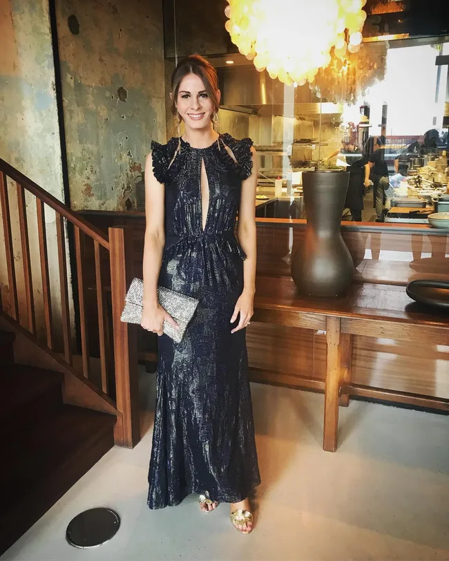 A woman in a dark blue evening gown holding a silver clutch, standing in a modern room with wooden decor.