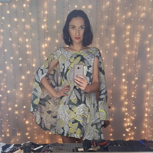 Person in a floral dress takes a mirror selfie against a backdrop of string lights.