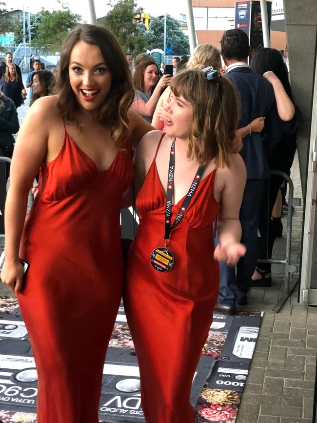 Two women in matching red dresses pose excitedly on a red carpet, surrounded by other attendees.