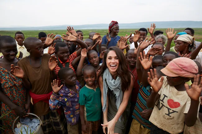 A woman smiling with a group of children and adults in a rural outdoor setting, many with hands raised in joy.