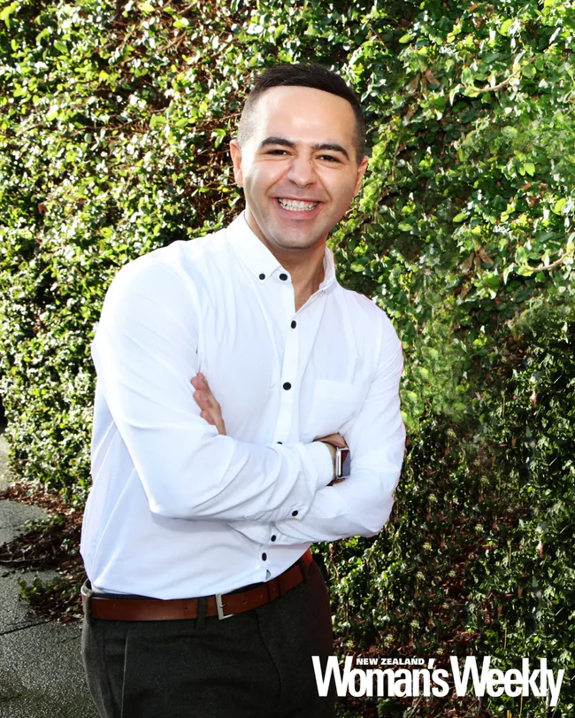Man in white shirt smiling with arms crossed, standing outside against a leafy green background.