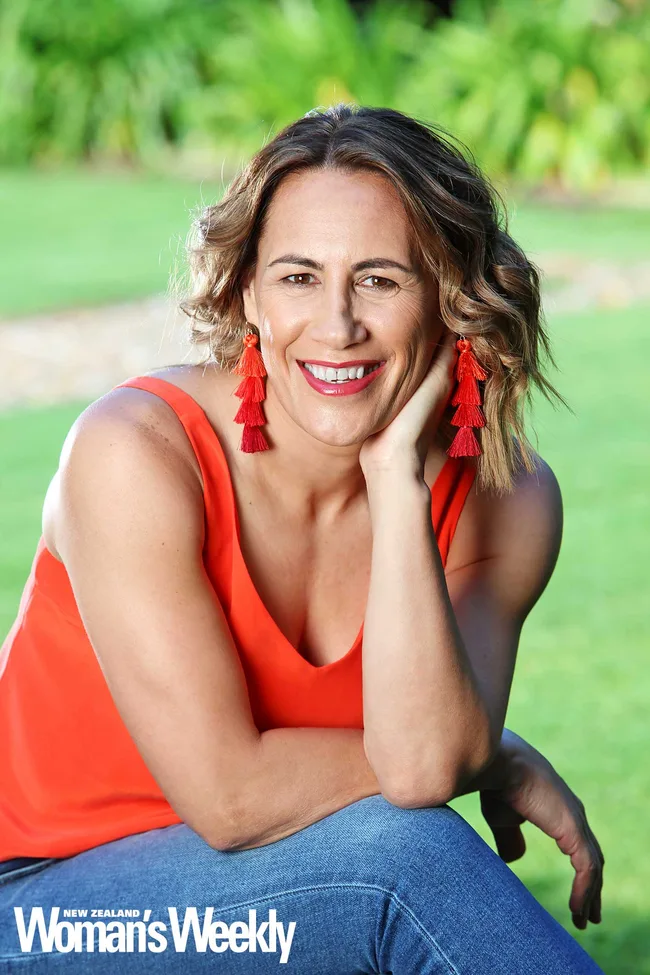 A woman in a red top and earrings smiles, sitting outdoors on grass.